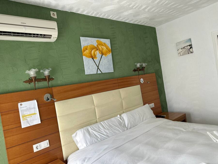 Una habitación de hotel con una pared verde, un cabecero de madera, una cama con ropa de cama blanca, un equipo de aire acondicionado y un cuadro de flores amarillas.