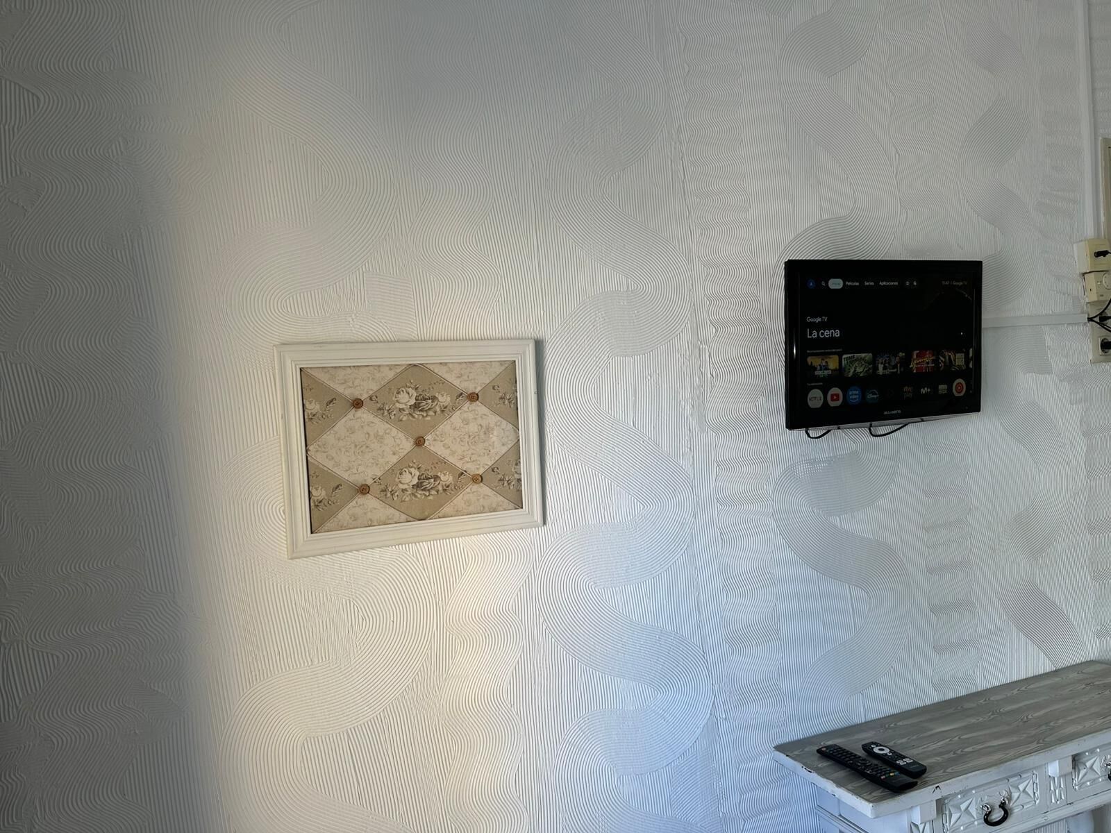 Een muur met een structuur, een ingelijst kunstwerk, een apparaat met iconen en een houten plank met afstandsbedieningen.