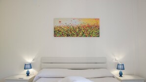 Une chambre à coucher avec un lit blanc, deux tables de chevet avec des lampes, une tête de lit blanche et un tableau au mur.