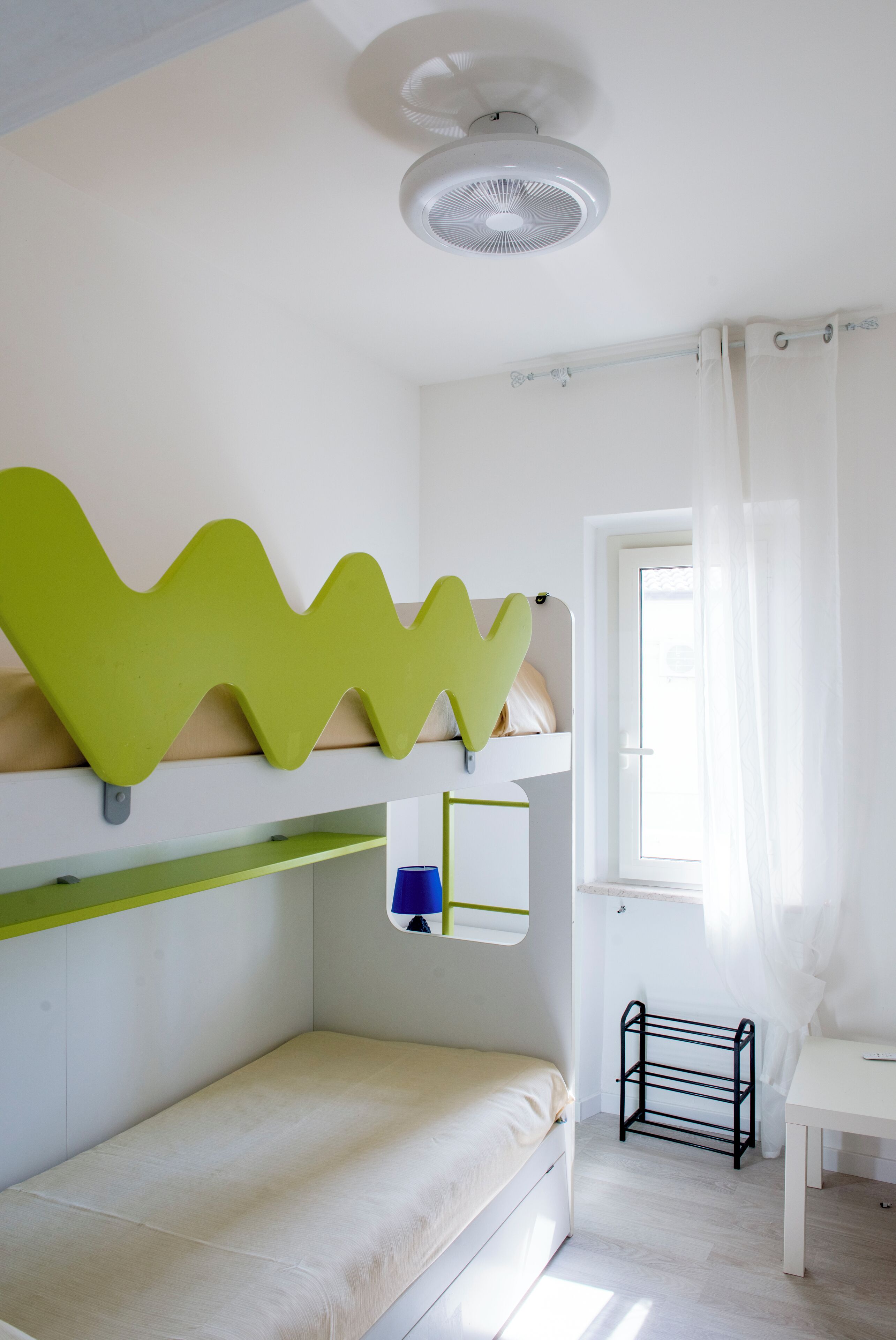 Una stanza con un letto a castello, una scrivania, una finestra con tende e una ventola a soffitto.