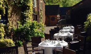 Outdoor dining area with tables and chairs set amidst a lush garden and brick building.
