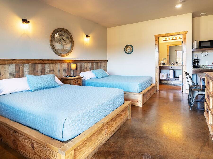 A room with two wooden beds, a wooden dresser, a kitchen area with a microwave, and a wall clock.
