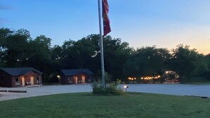 A grassy area with a flagpole, a body of water, and two buildings in the background. - The Woods Lodging (Madill)