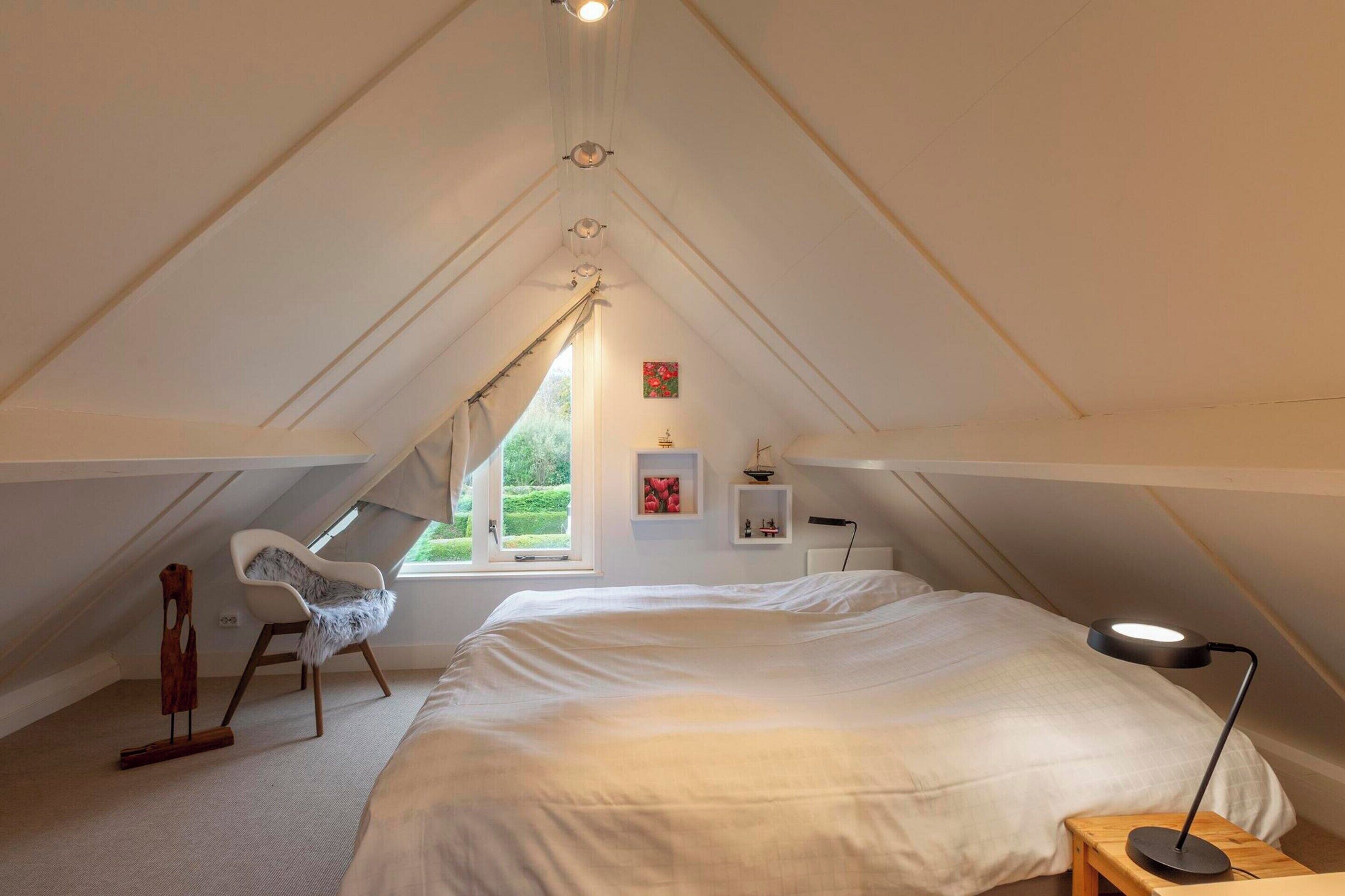Ein Schlafzimmer mit einem großen Bett, einem Sessel, einem Schreibtisch, einer Lampe und einem Fenster mit Blick auf Grünflächen.