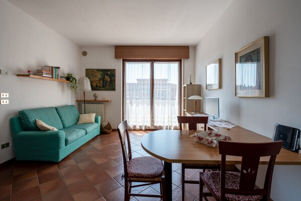 A living room with a teal sofa, a wooden dining table, and chairs. There is a window with sheer curtains, a lamp, and a bookshelf with books and a plant. - Apartment with garage near the center of Verona (Verona)