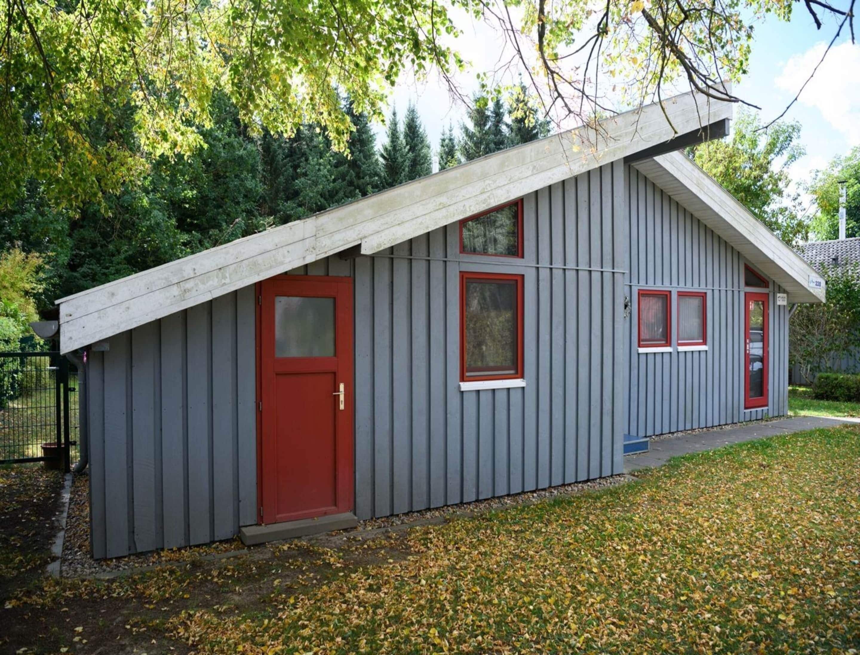 A small, single-story building with a red door and windows, surrounded by trees and a grassy area.