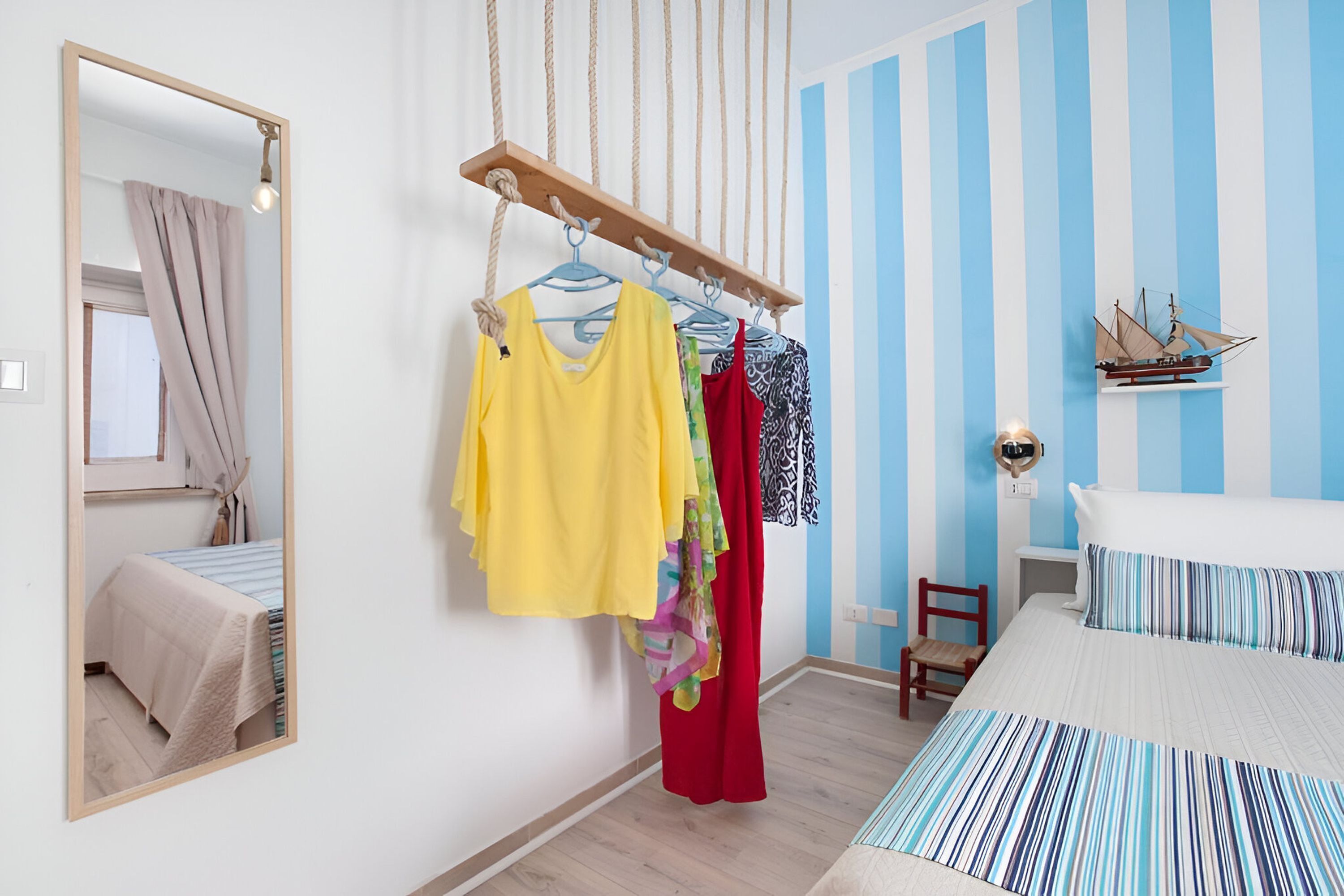A bedroom with a bed, a hanging clothes rack, a mirror, a small chair, and a ship model on the wall.