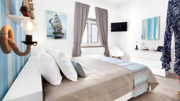 Un dormitorio con cama, cortinas, escritorio, silla, un cuadro de un barco y televisor montado en la pared.