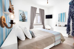 Un dormitorio con cama, cortinas, escritorio, silla, un cuadro de un barco y televisor montado en la pared.