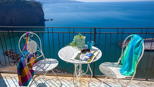 A balcony with a white metal railing, two white metal chairs, a small round table with a magazine, a blue mug, and a colorful bag.
