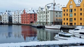 Une rangée de bâtiments aux couleurs vives longe le bord de l’eau, la neige recouvre le sol et des bateaux sont amarrés à proximité.