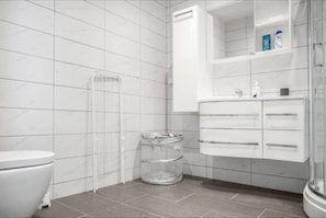 A bathroom with white tiled walls, a white toilet, a glass waste bin, and a white vanity with drawers. - Family Friendly, Free Parking Space, Short Walk to Brosundet (Ålesund)