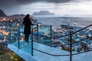 A person standing on a glass balcony overlooking a coastal town with buildings and a view of the sea. - Family Friendly, Free Parking Space, Short Walk to Brosundet (Ålesund)