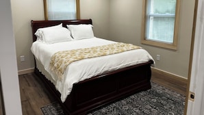 A bedroom with a wooden bed, white bedding, a patterned throw, two windows with blinds, a ceiling fan, and a dark patterned rug. - Cozy 2-bedroom cabin in charming Lexington with WiFi, AC (Lexington)