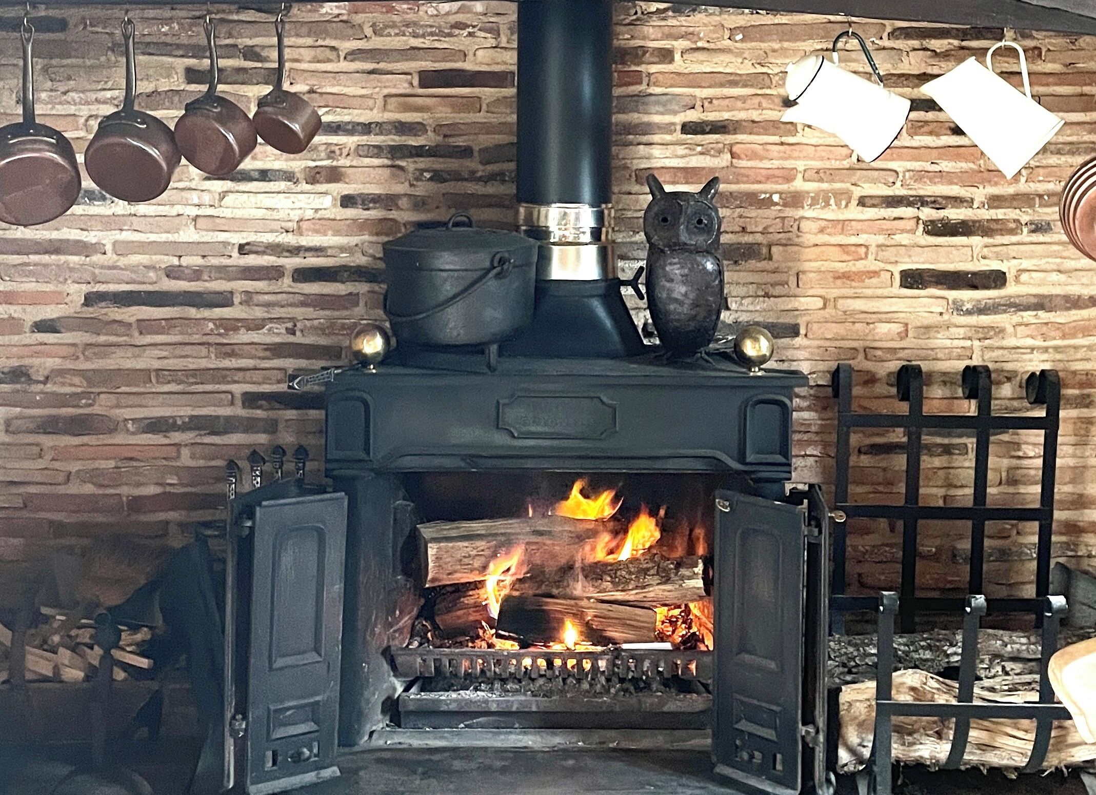 A wood-burning stove with pots and pans hanging above, set against a brick wall.