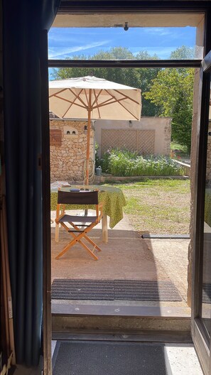 Vista a través de un ventanal a un patio con mesa y sombrilla, rodeado de follaje y un muro de piedra.