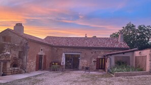 Un edificio rústico de piedra con tejado de baldosas, una entrada central y un pequeño patio con mesa y sillas.