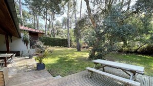 Una terraza de madera con una mesa y sillas de picnic, rodeada de un jardín exuberante y altos árboles.