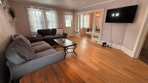 A living room with a sectional sofa, a wooden coffee table, a flat-screen TV mounted on the wall, and a dining area with a table and chairs.