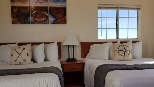 A hotel room with two beds, a nightstand with a lamp, a window with blinds, and a wall with a compass-themed pillow.