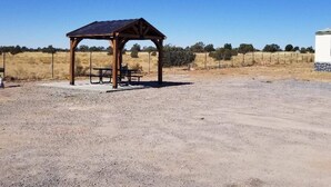 A covered outdoor area with a picnic table and benches, surrounded by a gravel ground and sparse vegetation.