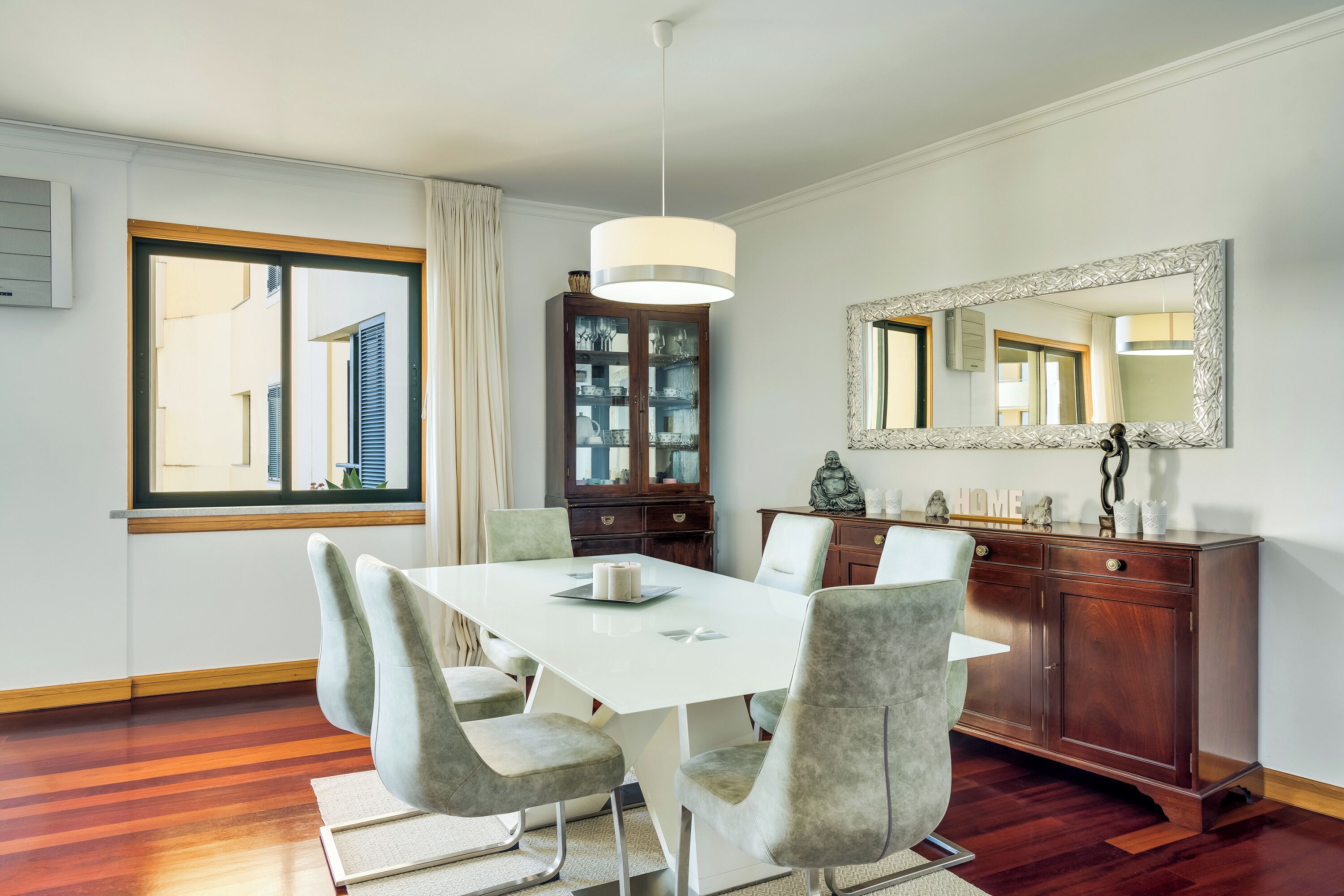 A dining area with a white table, grey upholstered chairs, a wooden cabinet, and a large mirror on the wall.