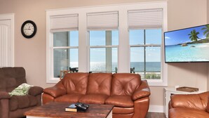 A living room with a brown leather sofa, a wooden coffee table with books and a remote control, a TV displaying a beach scene, and large windows with blinds. - Windchimes (Port St. Joe)