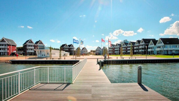 A waterfront hotel with a wooden pier, multiple buildings with balconies, and flags. - Ferienhaus im Ferienpark Mueritz (Rechlin)