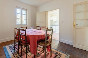A dining area with a round table, six wooden chairs, and a red tablecloth. There is a window with a view of trees and a door leading to another room. - La Maison Vergès - 3 bedrooms - large living room - small garden (Ossun)