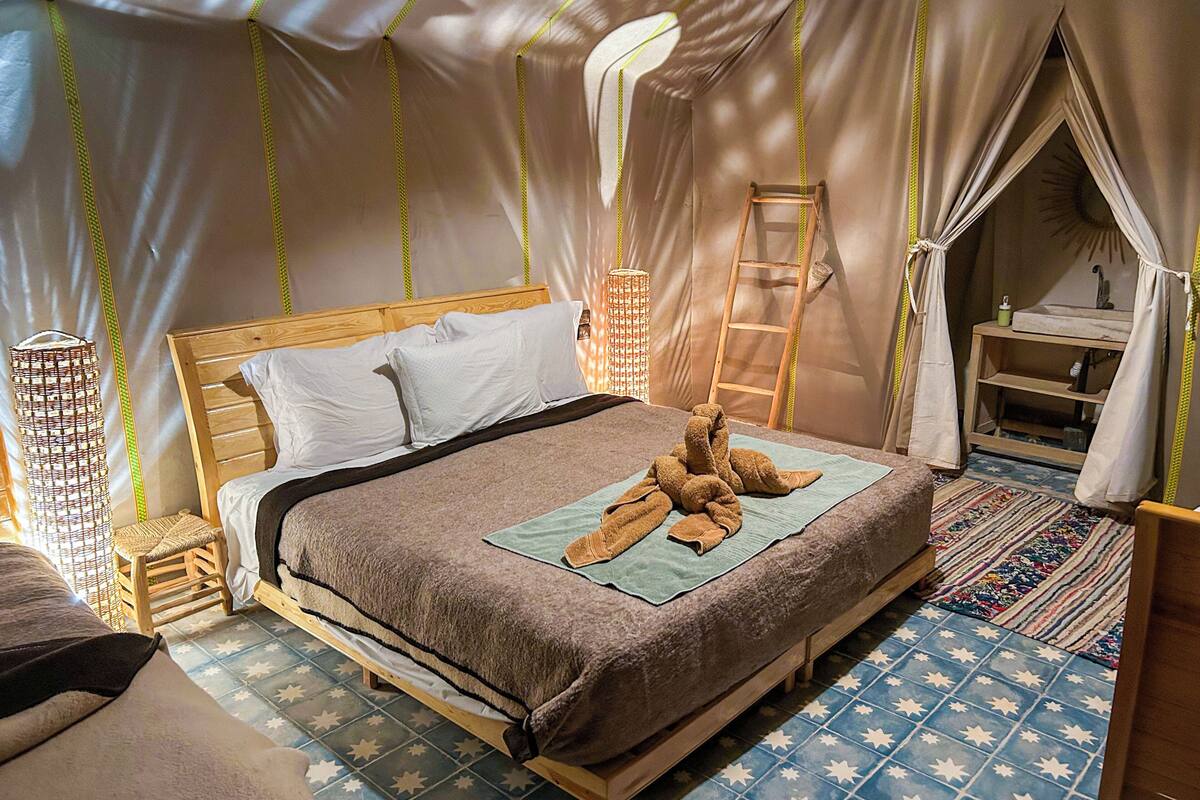 Un dormitorio estilo tienda de campaña, con una cama de madera, una escalera, un lavamanos pequeño y una lámpara de mimbre.