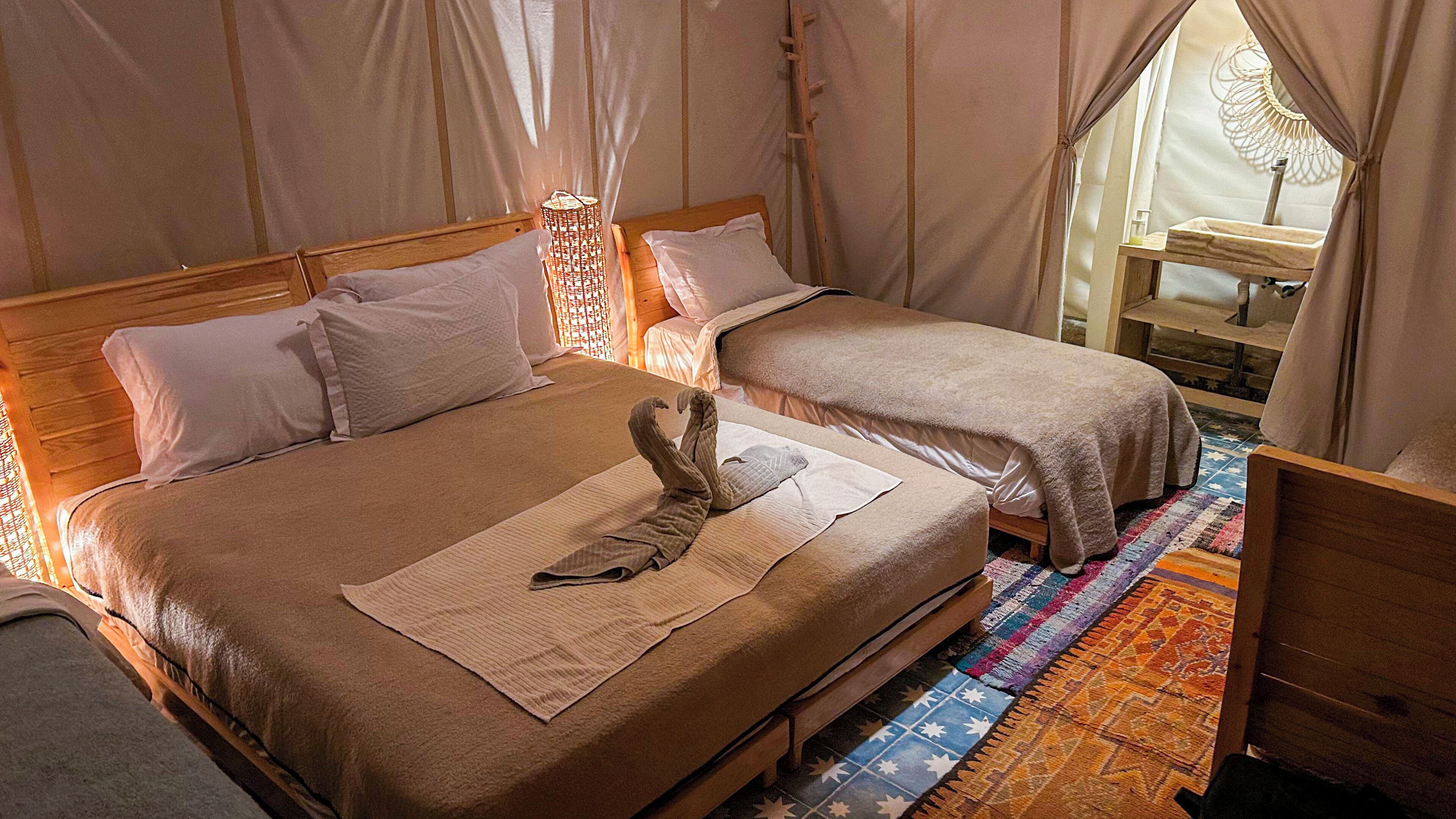Een kamer met twee bedden, een houten hoofdbord, een geweven hemelbed, een kamerkleed met patroon en een houten nachtkastje.