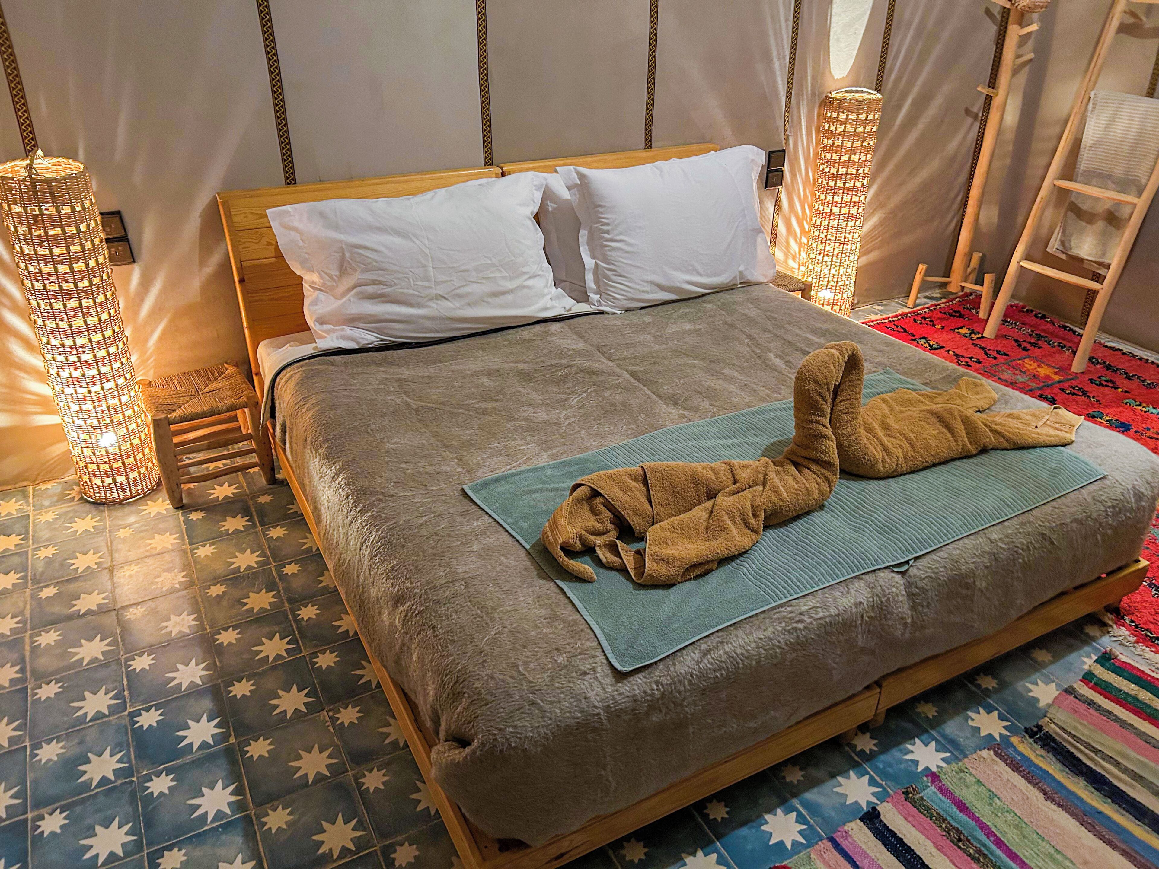 Una cama con una manta marrón y una toalla azul, una lámpara de mimbre y un suelo con estampado de estrellas.