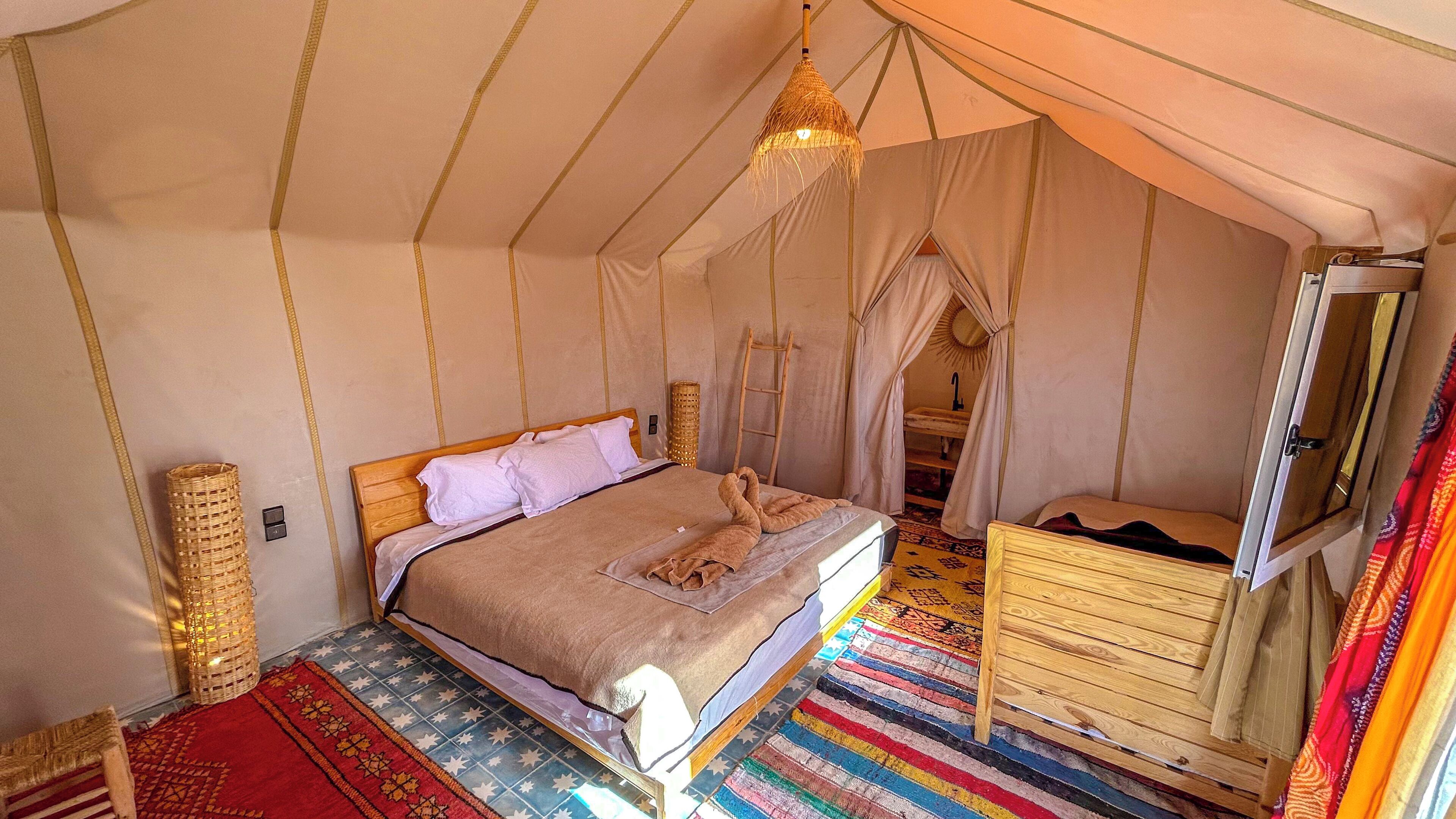 Een tentachtige kamer met een bed, een houten dressoir, een hanglamp en een kleurrijk gestreept vloerkleed.