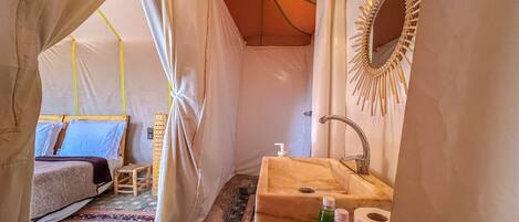 Habitación tipo tienda de campaña con una cama, un taburete pequeño, un lavabo y un espejo.