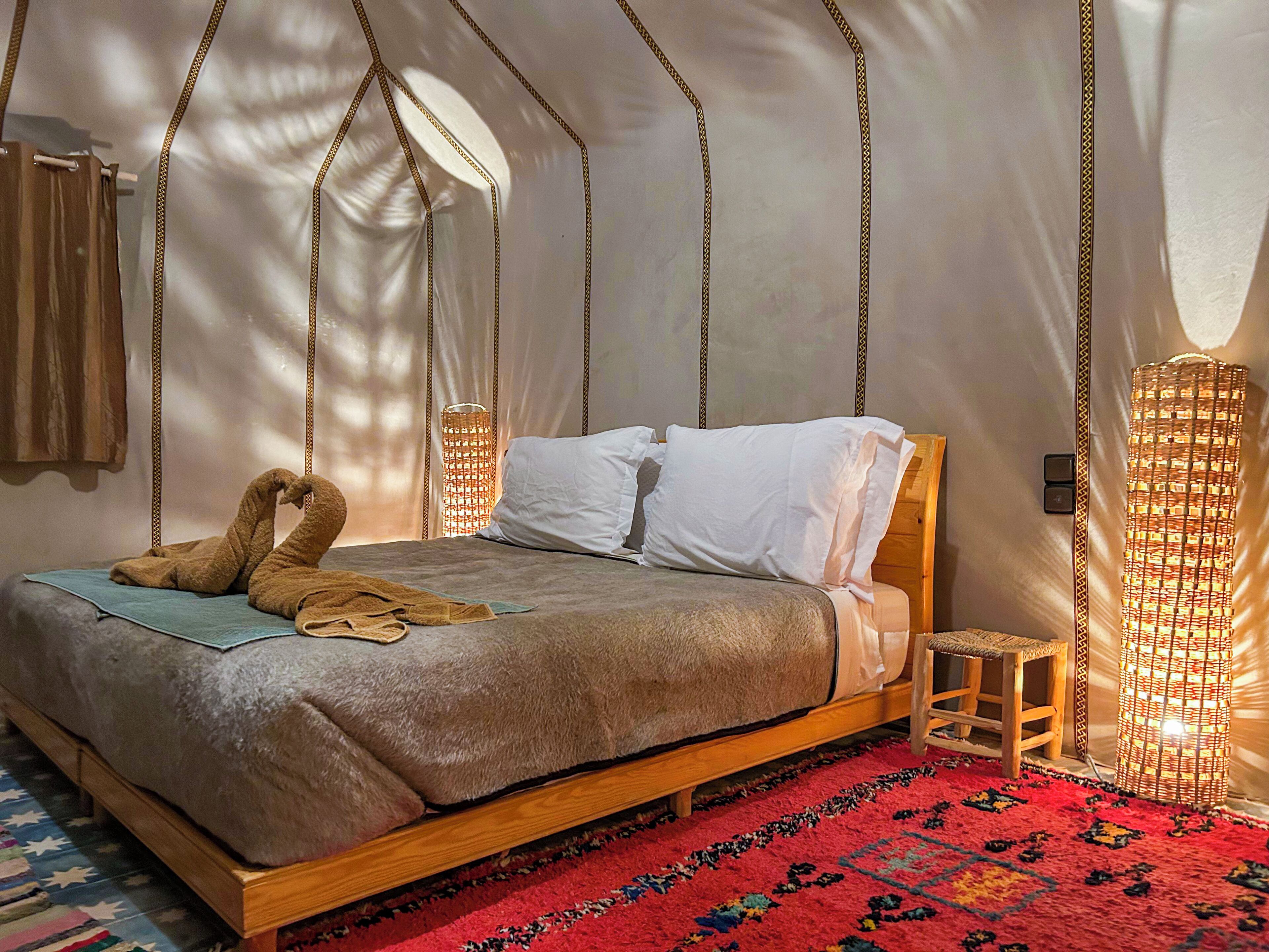 Una cama con estructura de madera, almohadas blancas y una manta marrón en una habitación tipo tienda con alfombra roja.