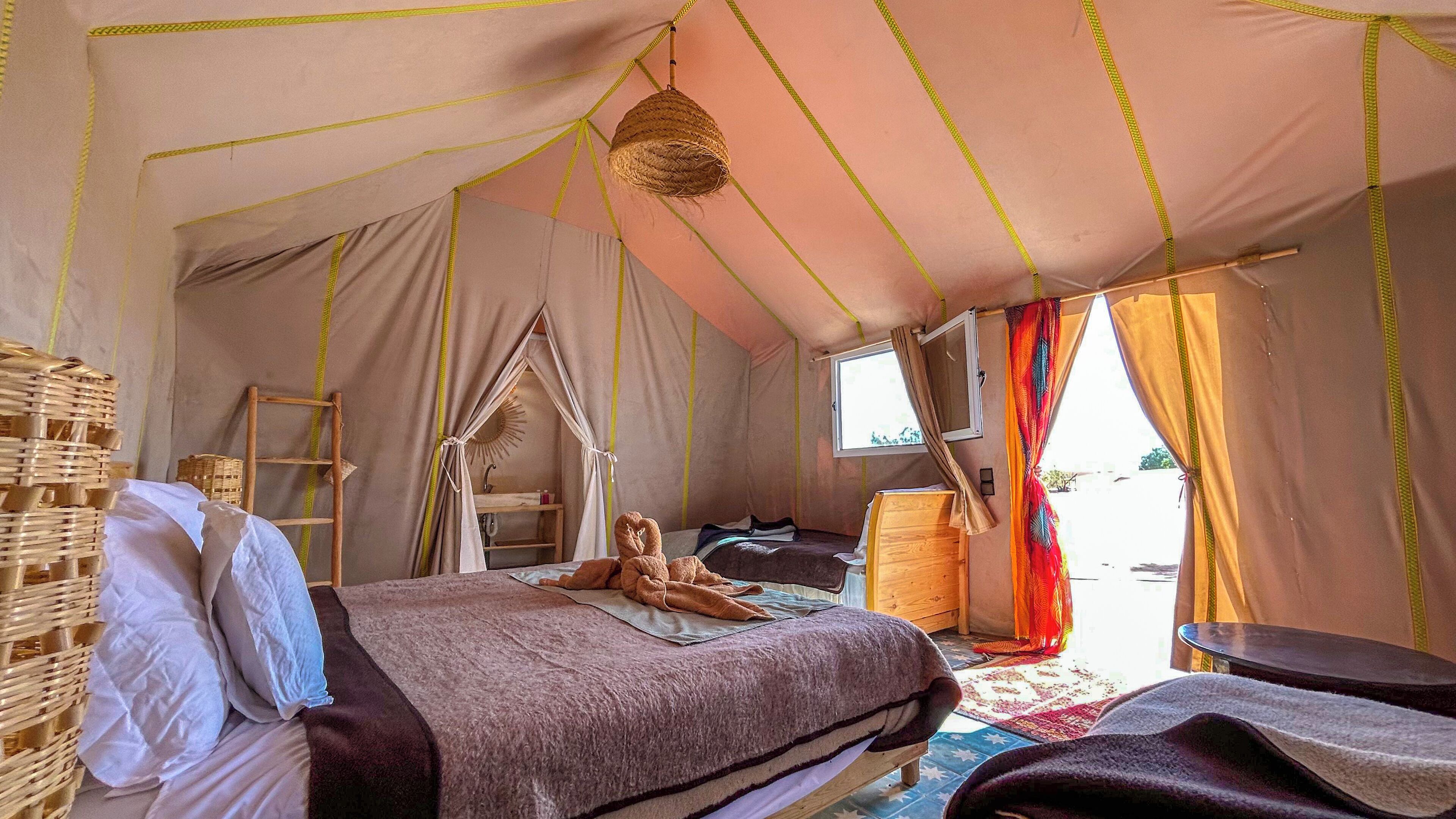 A tented room with a bed, a small table, a chair, a hanging basket, and a window.
