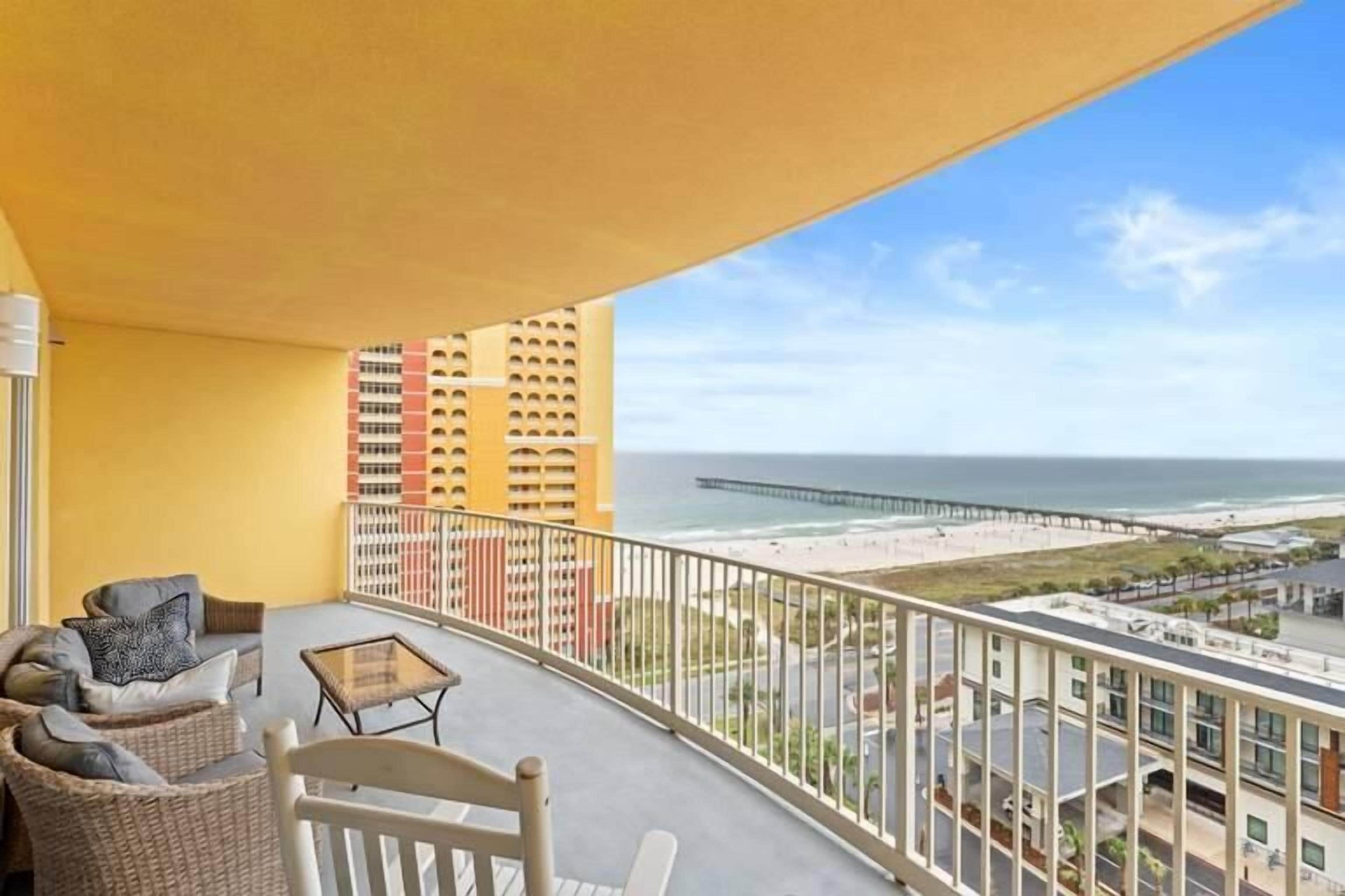 Ein Balkon mit Korbmöbeln und einem Glastisch mit Blick auf einen Strand und einen Steg.
