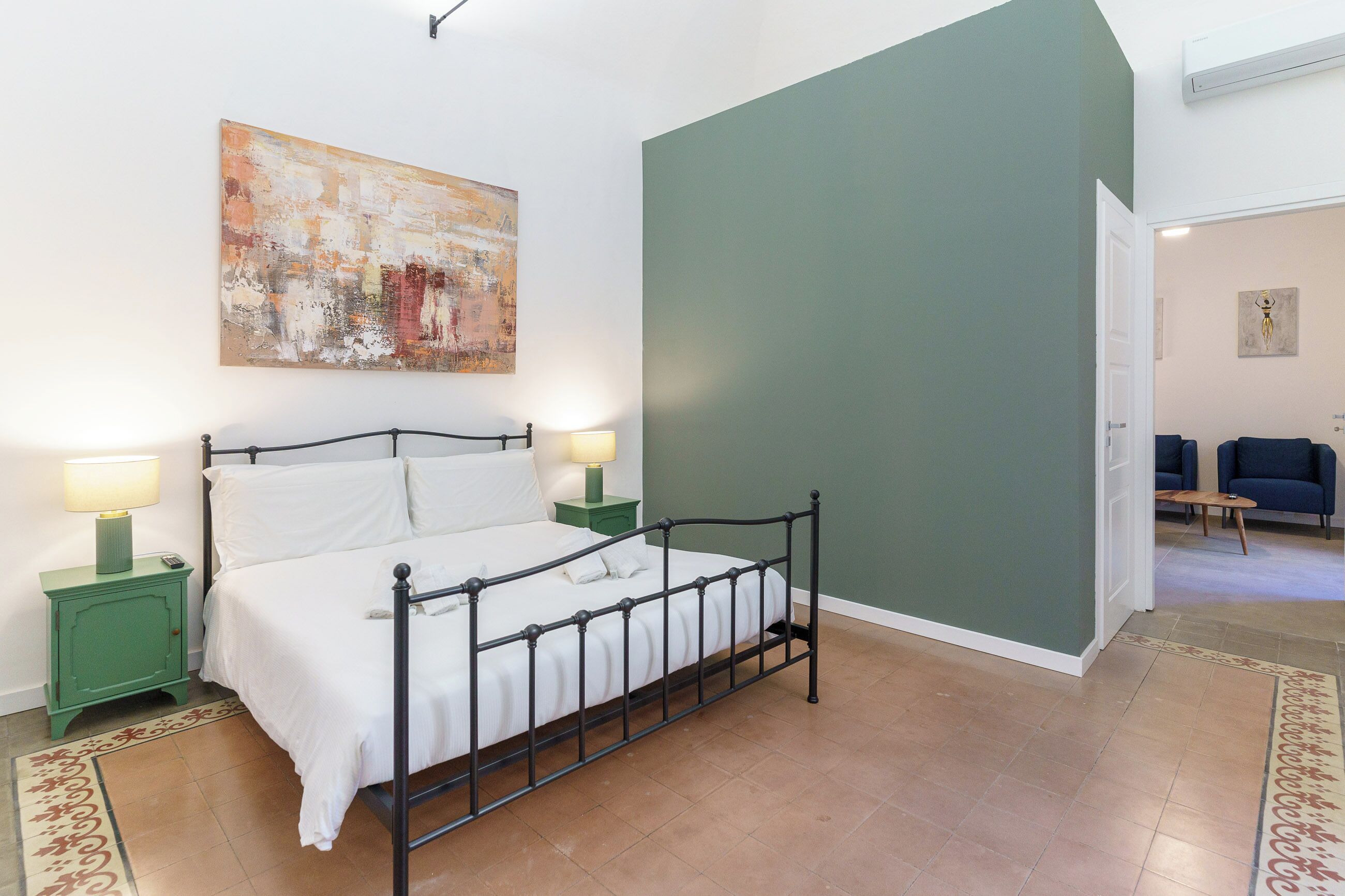 A bedroom with a metal bed frame, two bedside tables with lamps, a green wall, and a large abstract painting.