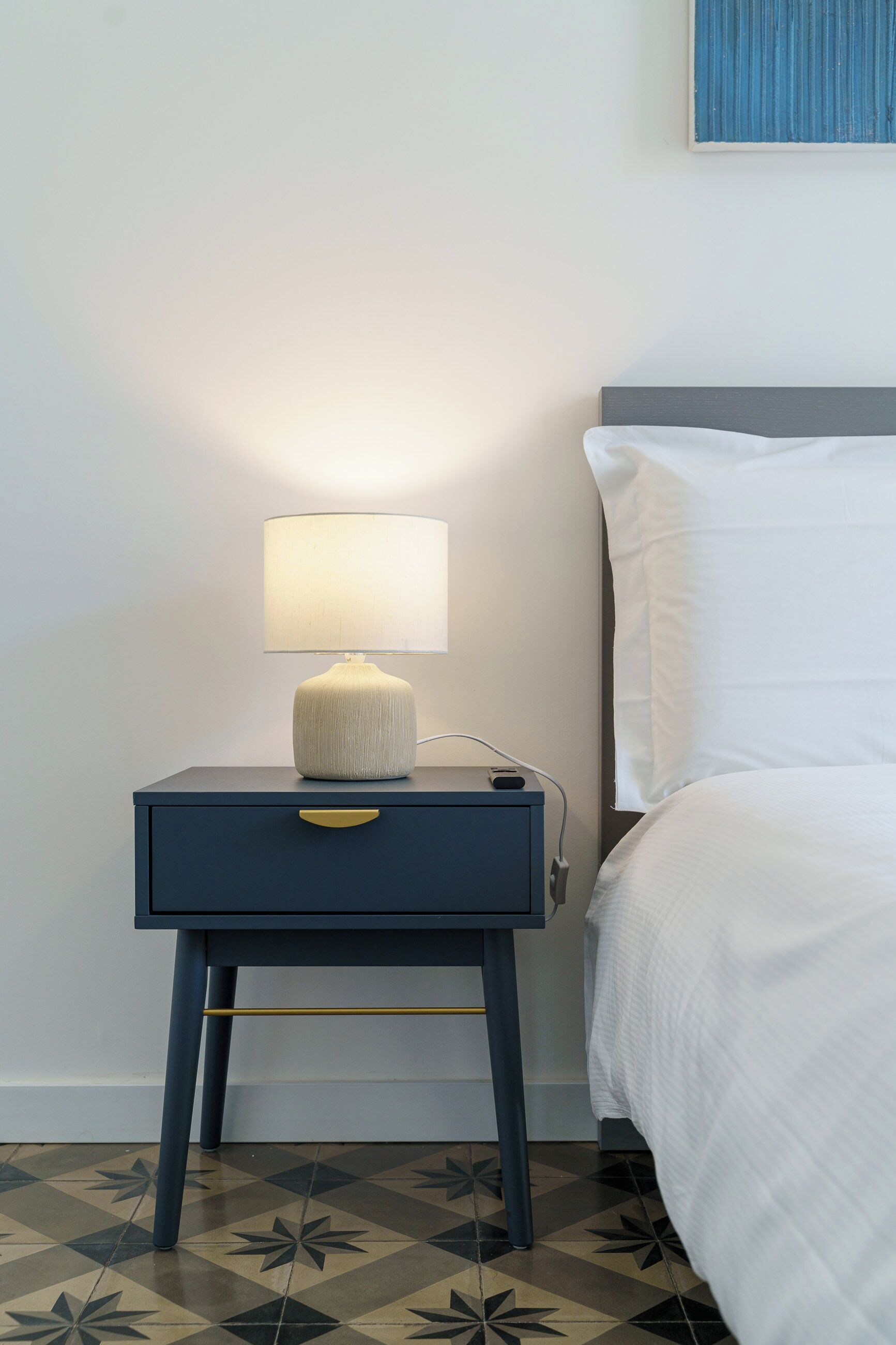 A neatly made bed with white bedding, a bedside table with a lamp, and a patterned floor.