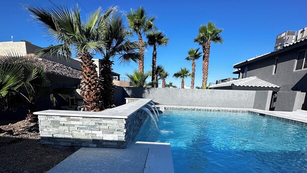 A swimming pool with a stone border, palm trees, and a clear blue sky.