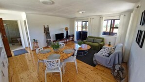 Un vaste espace de vie doté d’un plancher en bois, d’une table de salle à manger ronde entourée de chaises blanches, d’un canapé vert et d’un fauteuil gris.