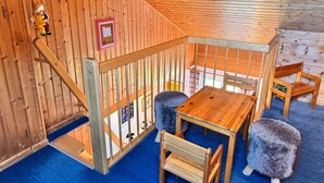 A wooden cabin interior with a wooden table, two chairs, a small stool, a swing, and a framed picture on the wall.
