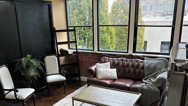 Ein Wohnzimmer mit Ledersofa, zwei Sesseln, einem Couchtisch und einem großen Fenster mit Blick auf Bäume und Gebäude.
