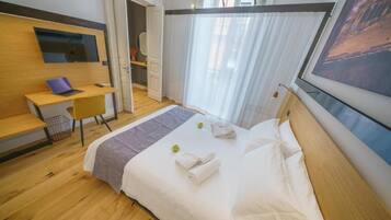 Een moderne hotelkamer met een houten vloer, een bed met witte lakens, een bureau met een laptop, een aan de muur bevestigde televisie en een groot raam met dunne gordijnen.