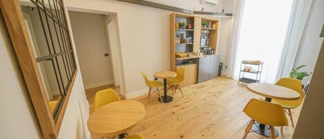 A modern interior with wooden flooring, yellow chairs, a small round table, and a kitchen area with shelves and a refrigerator.
