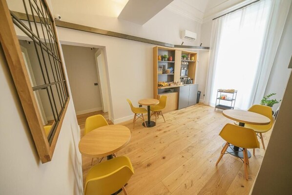 Un intérieur moderne avec un plancher en bois, des chaises jaunes, une petite table ronde et un coin cuisine doté d’étagères et d’un réfrigérateur.