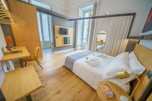 Una moderna camera d'albergo con pavimento in legno, un letto con lenzuola bianche, una scrivania con sedia, una TV montata a parete e un'ampia finestra con tende.