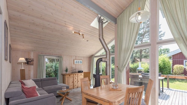 A cozy living room with a sofa, a dining table, and chairs. There is a kitchen area with cabinets and a stove. Large windows allow natural light and views of the outdoors. - Ferienhaus mit Sauna im Ferienpark Mirow (Mirow)