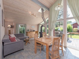 A cozy living room with a sofa, a dining table, and chairs. There is a kitchen area with cabinets and a stove. Large windows allow natural light and views of the outdoors.