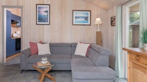 A living room with a grey sectional sofa, a glass coffee table, and a wooden cabinet. There are two framed pictures on the wall and a lamp beside the window. - Ferienhaus mit Sauna im Ferienpark Mirow (Mirow)